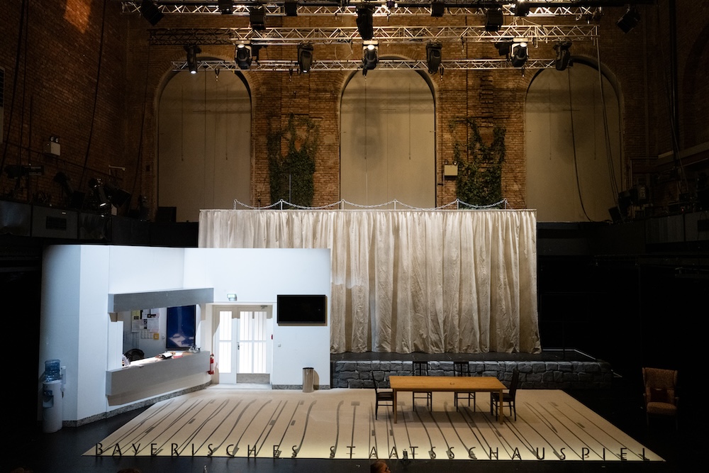 Stage design for Mitläufer at Residenztheater consisting of a set of the theater's entrance, a platform with a curtain, a table with four chairs. On the floor a line of letters forming BAYRISCHES STAATSSCHAUSPIEL and casting shadows on the floor.