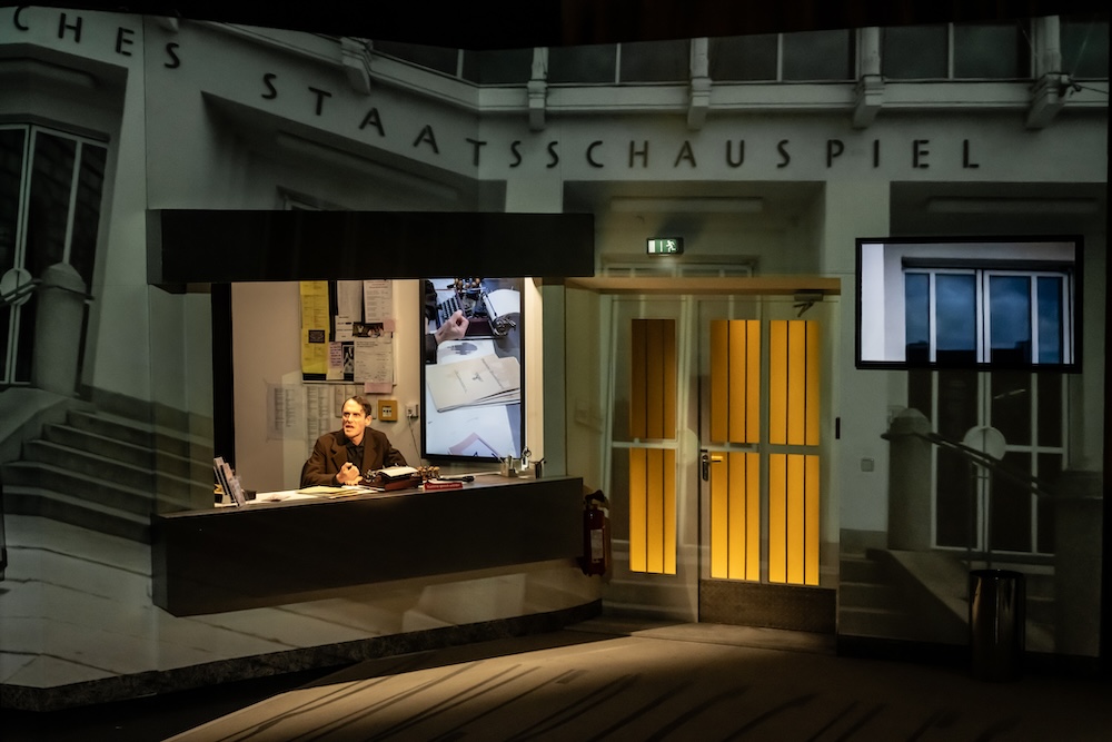 An actor on stage sitting in a set that looks like a theater entrance