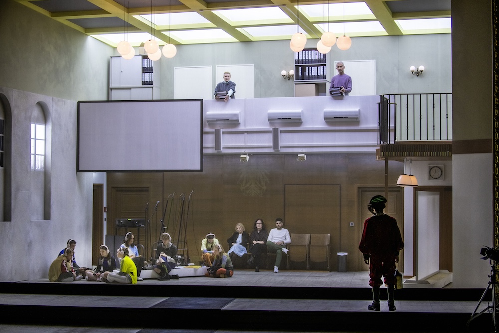 Total view of the stage of Nicht Sehen: An emtied out court room with a second floor balkony. Teenagers sitting on the floor, two people hold paperwork on the upper floor, one guard figure is watching it all from front of stage.