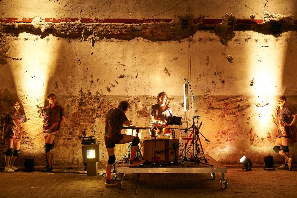 Two musicians on drumset and synthie in front of the ouside wall of a building.